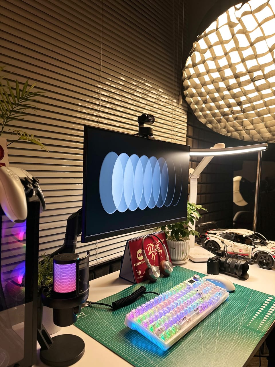 Setup góc máy của Drake nè: Góc bàn tối ánh vàng với bàn phím RGB trong suốt, micro hồng tím, đèn bàn, cây xanh và mô hình xe Lego Porsche.