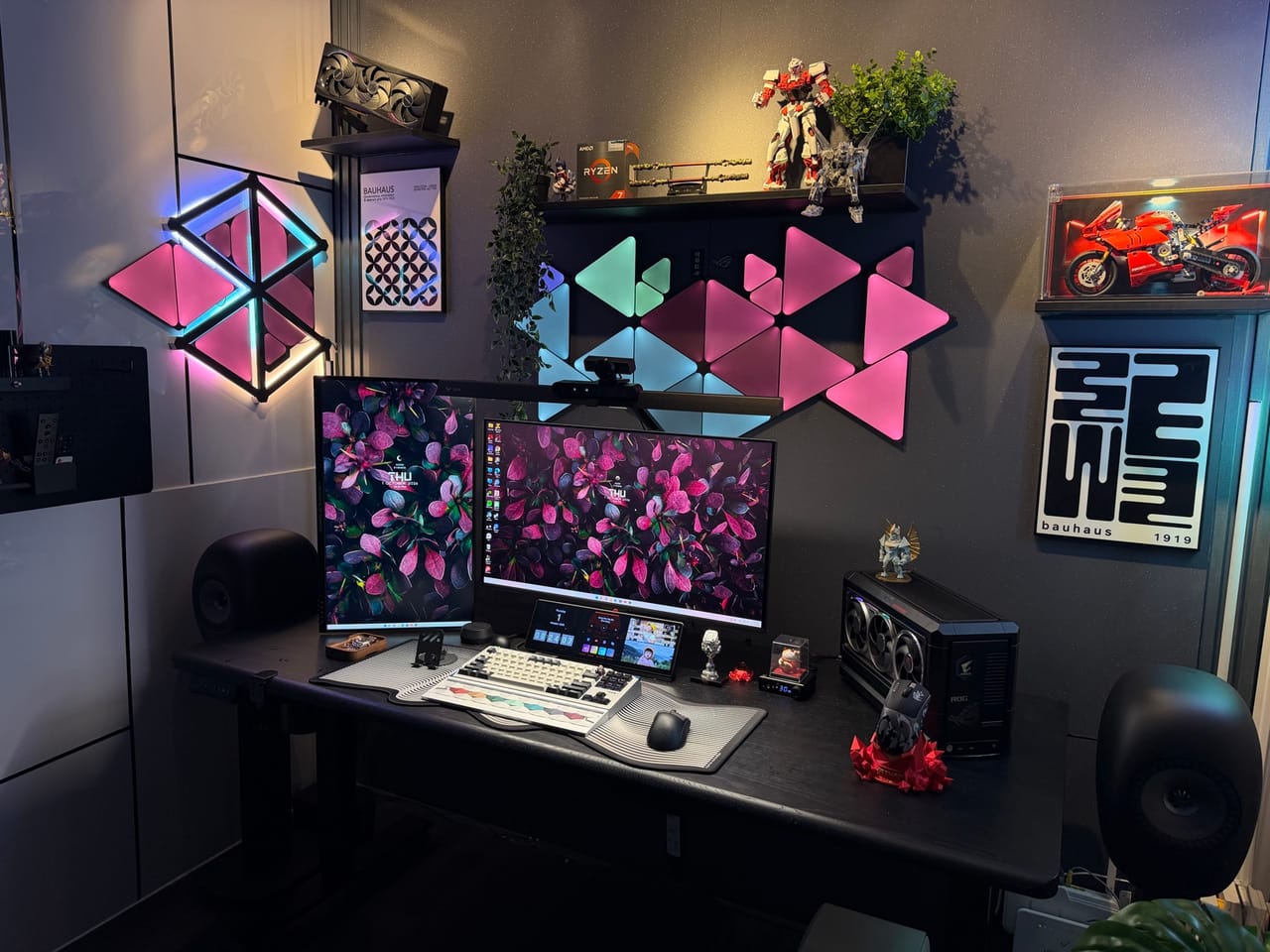 Góc máy của Thanh Duy: Góc setup tông đen với đèn panel RGB hồng và xanh trên tường, hai màn hình, loa, mô hình anime, tranh Bauhaus và cây xanh trang trí.