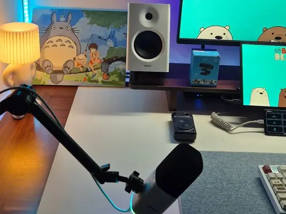 Góc máy của Khánh99: Góc bàn trắng với đèn LED xanh dương, tranh Totoro, loa trắng, micro có arm, hai màn hình hiển thị hình We Bare Bears và bàn phím cơ.