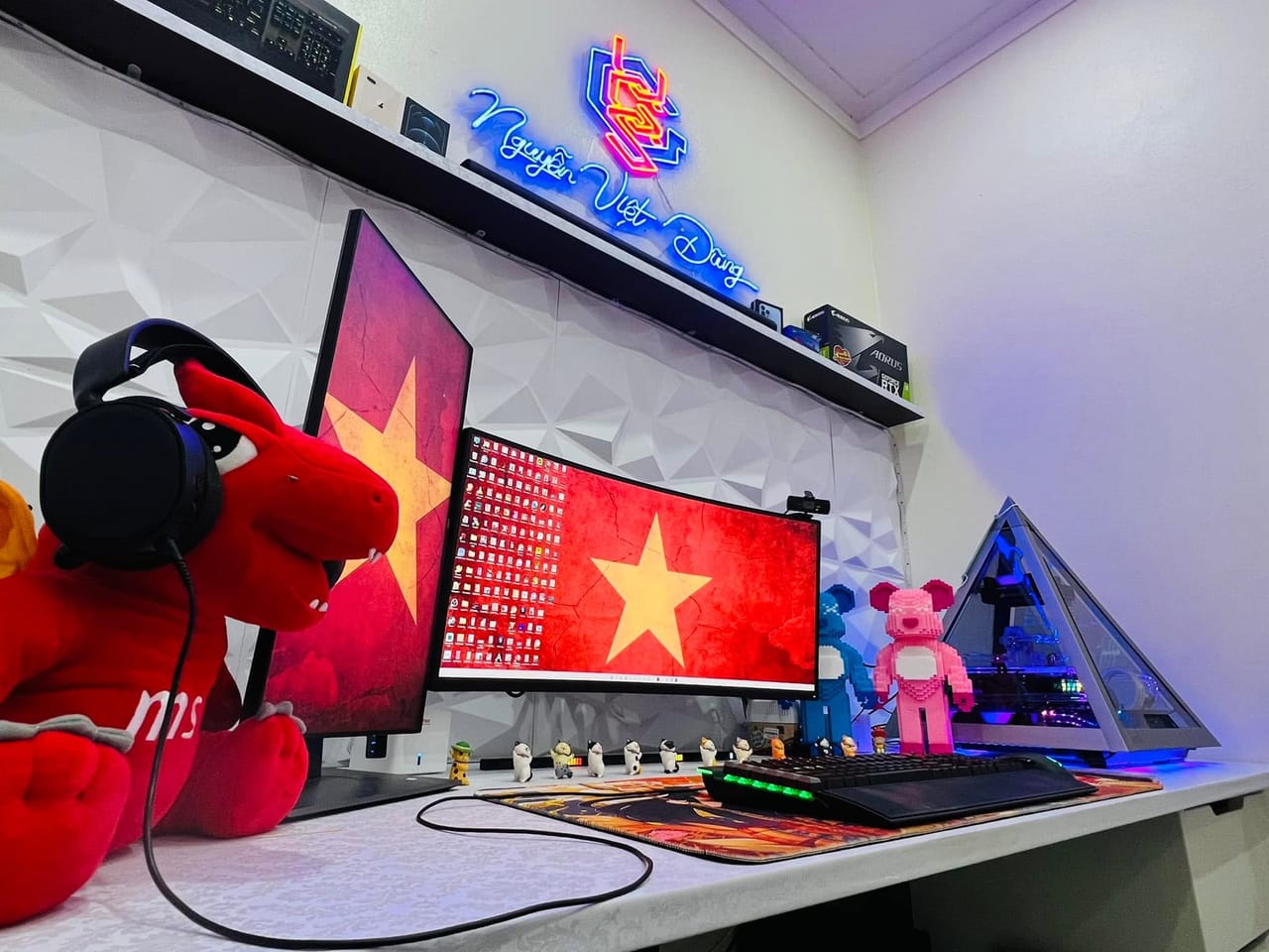 Góc máy của Nguyễn Việt Dũng: Góc setup nền tường trắng 3D với hai màn hình hình cờ đỏ sao vàng, case kim tự tháp, đèn neon và mô hình gấu.