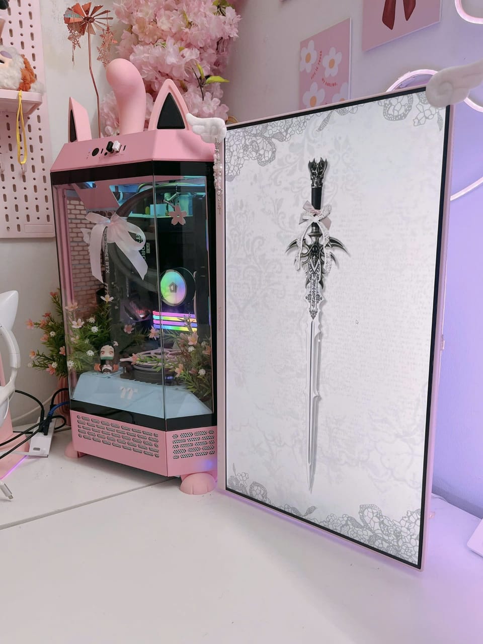 Góc máy của Dánh Gaming: Góc setup tông hồng pastel với case tai mèo trang trí hoa và figure anime, bên cạnh màn hình dựng dọc hình thanh kiếm nền ren.