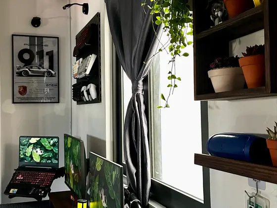 Góc máy của Võ Minh Sang: Góc setup nhỏ cạnh cửa sổ với laptop, hai màn hình hình nền lá xanh, hai bàn phím, kệ gỗ và cây cảnh.