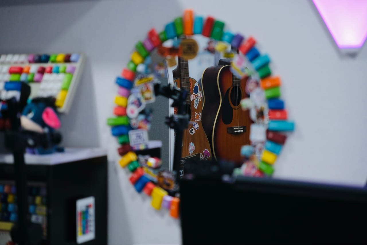 Góc máy của Thư Đoàn: Góc setup với gương viền keycap nhiều màu phản chiếu cây đàn guitar dán sticker, cạnh đèn LED tím hồng và bàn phím custom.