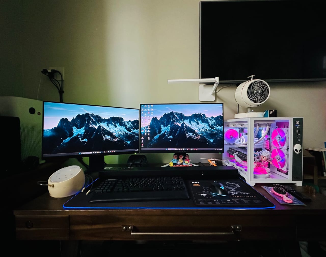 Góc máy của Thảo Thông Thạo: Góc máy với hai màn hình, case trắng đèn hồng RGB, bàn phím và chuột trên bàn gỗ, cùng tivi treo tường phía sau.