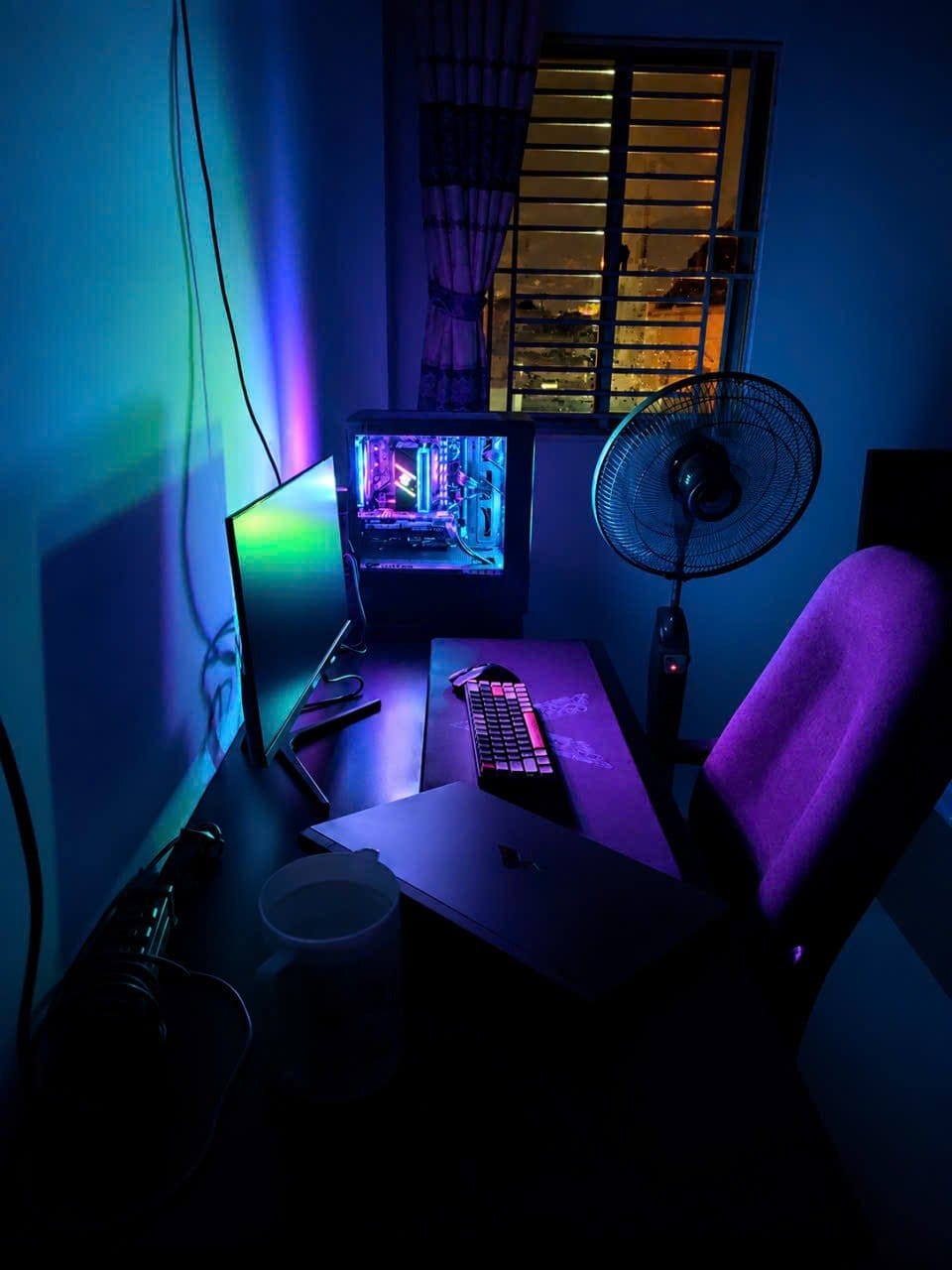 Góc máy của DoreNguyên: Góc máy ban đêm ngập ánh đèn RGB xanh tím, có PC case kính, bàn phím cơ, laptop, ghế tím và quạt đứng cạnh cửa sổ.