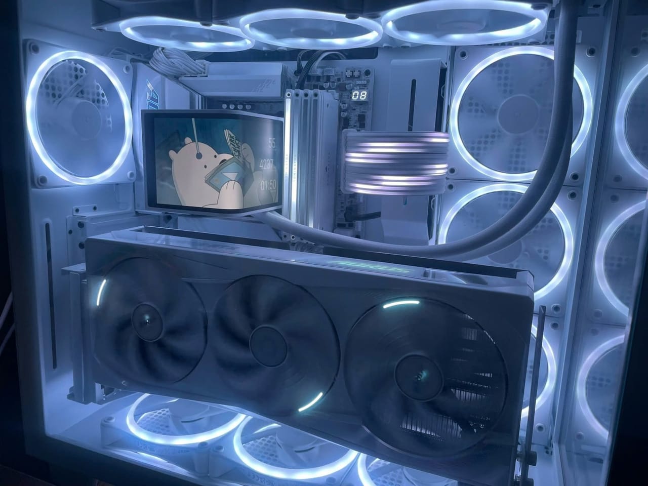 Góc máy của S1zz: Bên trong case PC kính cường lực màu trắng với nhiều quạt LED sáng xanh, card đồ họa AORUS và tản nhiệt nước có màn hình nhỏ hiển thị hình hoạt hình.