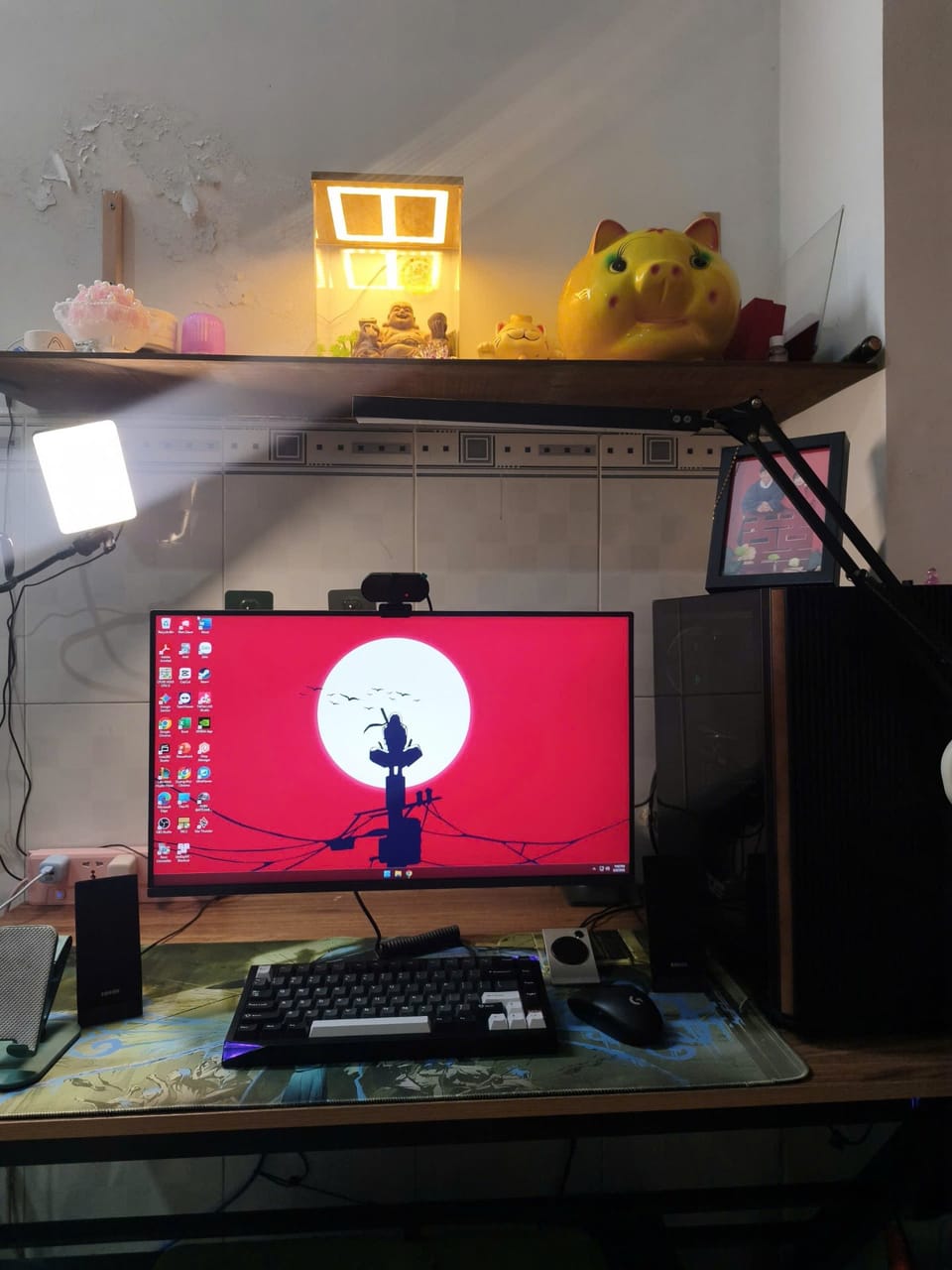 Setup góc máy của QuangPhu: Góc bàn làm việc với một màn hình hình nền anime đỏ, bàn phím cơ, chuột đen, loa, webcam, đèn livestream và kệ trang trí phía trên.