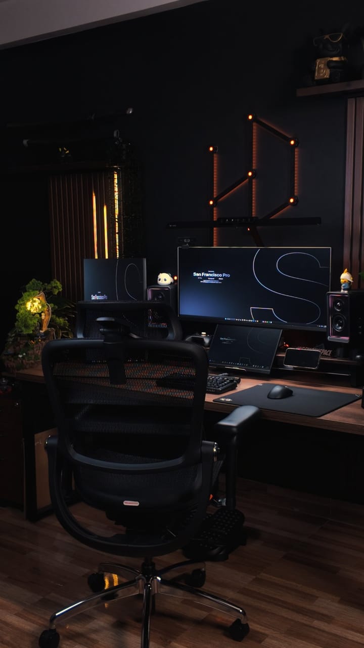 Setup góc máy của hdanggworkspace: Góc máy tối giản tông đen với bàn gỗ, màn hình rộng, loa, đèn LED cam trên tường, ghế công thái học và chậu cây nhỏ.