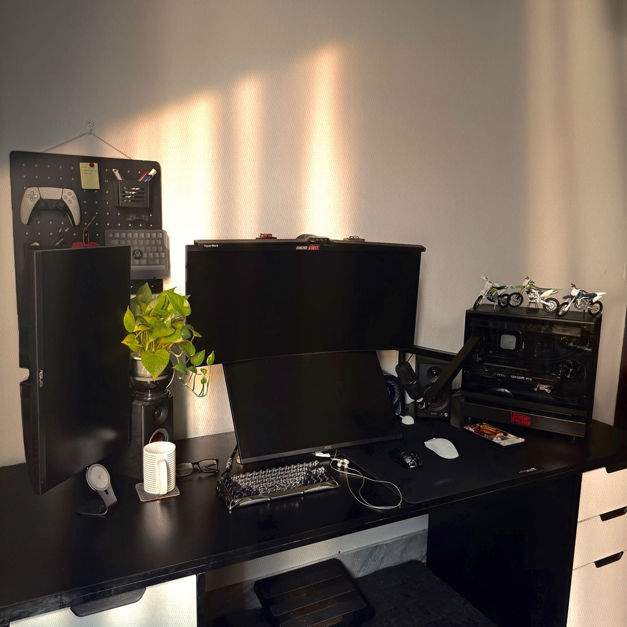 Setup góc máy của heat1e: Góc bàn tông đen với ba màn hình, pegboard treo tay cầm, chậu cây xanh và mô hình xe mô tô trên case PC, nắng chiều rọi tường.
