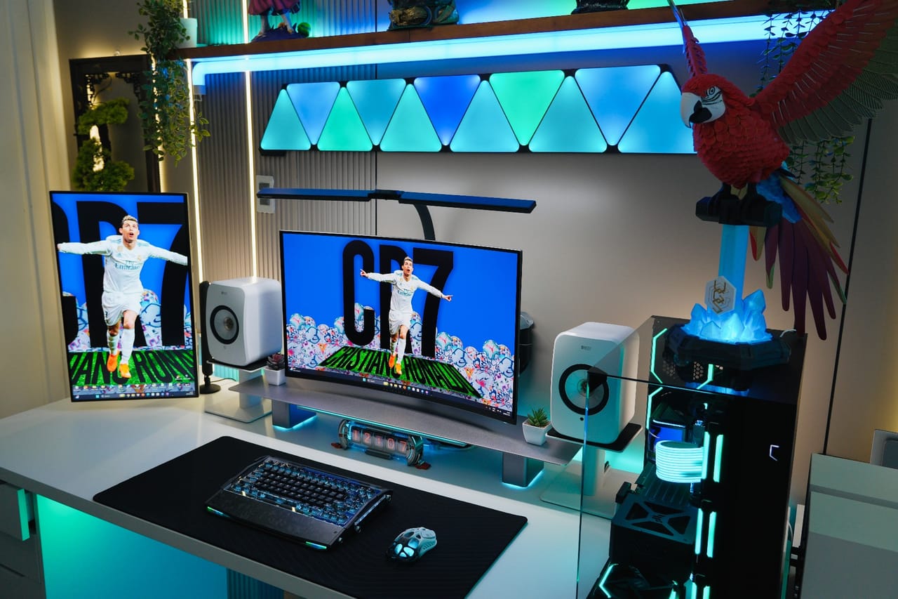Góc máy của Steven Huy: Góc setup tông trắng đen với đèn RGB xanh dương, hai màn hình, loa KEF, case kính, tượng vẹt đỏ và cây xanh trang trí.
