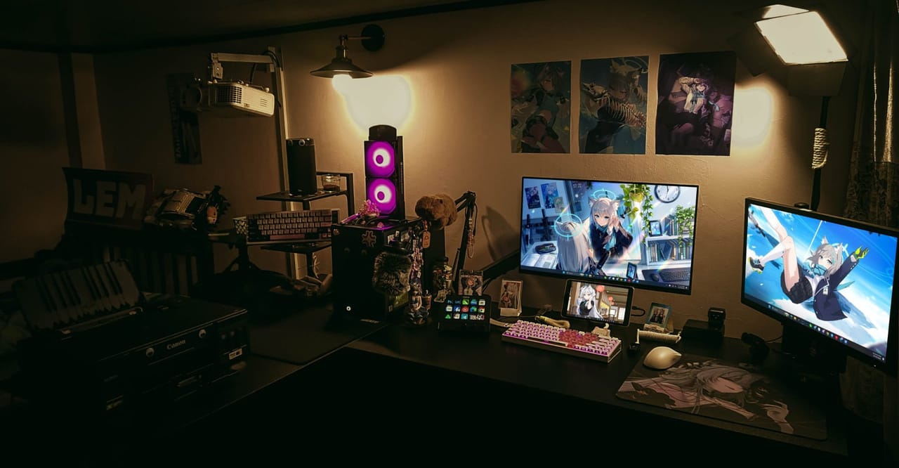 Góc máy của Tùng Lem: Góc setup tối ánh đèn vàng, bàn đen với hai màn hình hình nền anime, bàn phím hồng, case RGB tím, poster và máy chiếu trên tường.