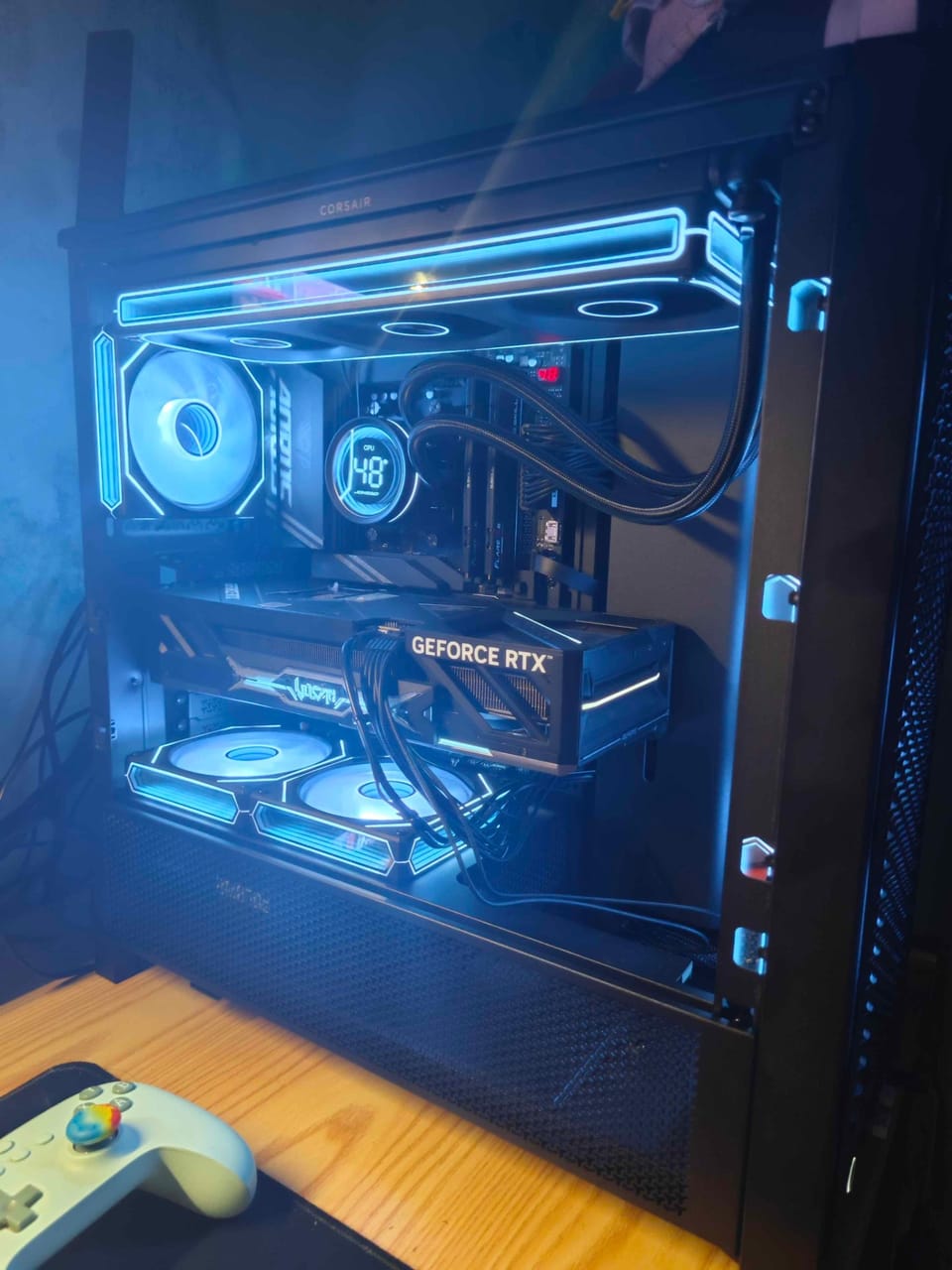 Góc máy của Apurusensei: Dàn PC case kính đen với quạt và viền LED xanh neon, card RTX, tản nhiệt nước hiển thị nhiệt độ CPU, đặt trên bàn gỗ cạnh tay cầm.