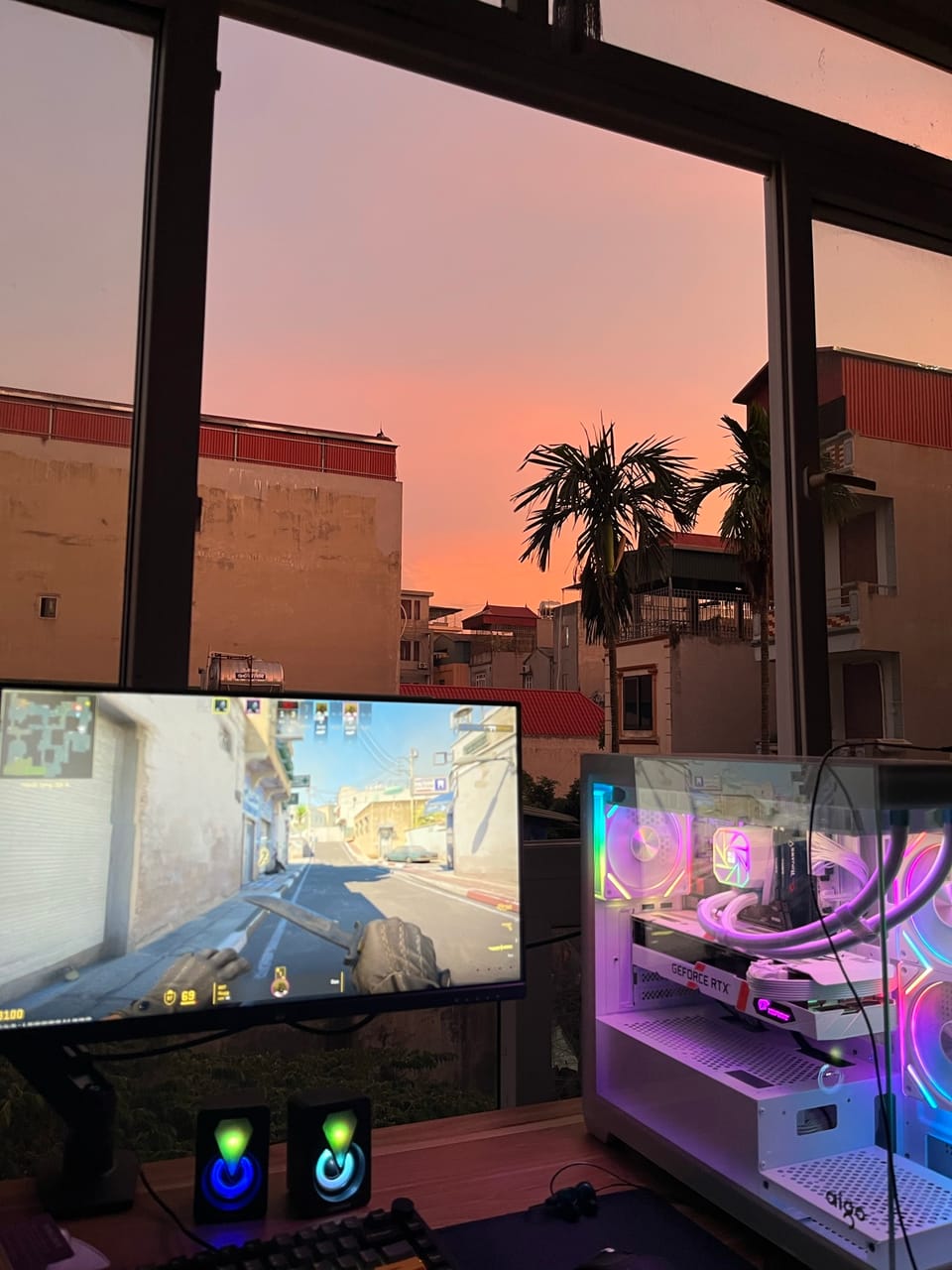 Góc máy của aug shadcn: Góc máy cạnh cửa sổ hoàng hôn hồng, case Aigo trắng RGB, card GeForce RTX, màn hình đang chơi CS2.