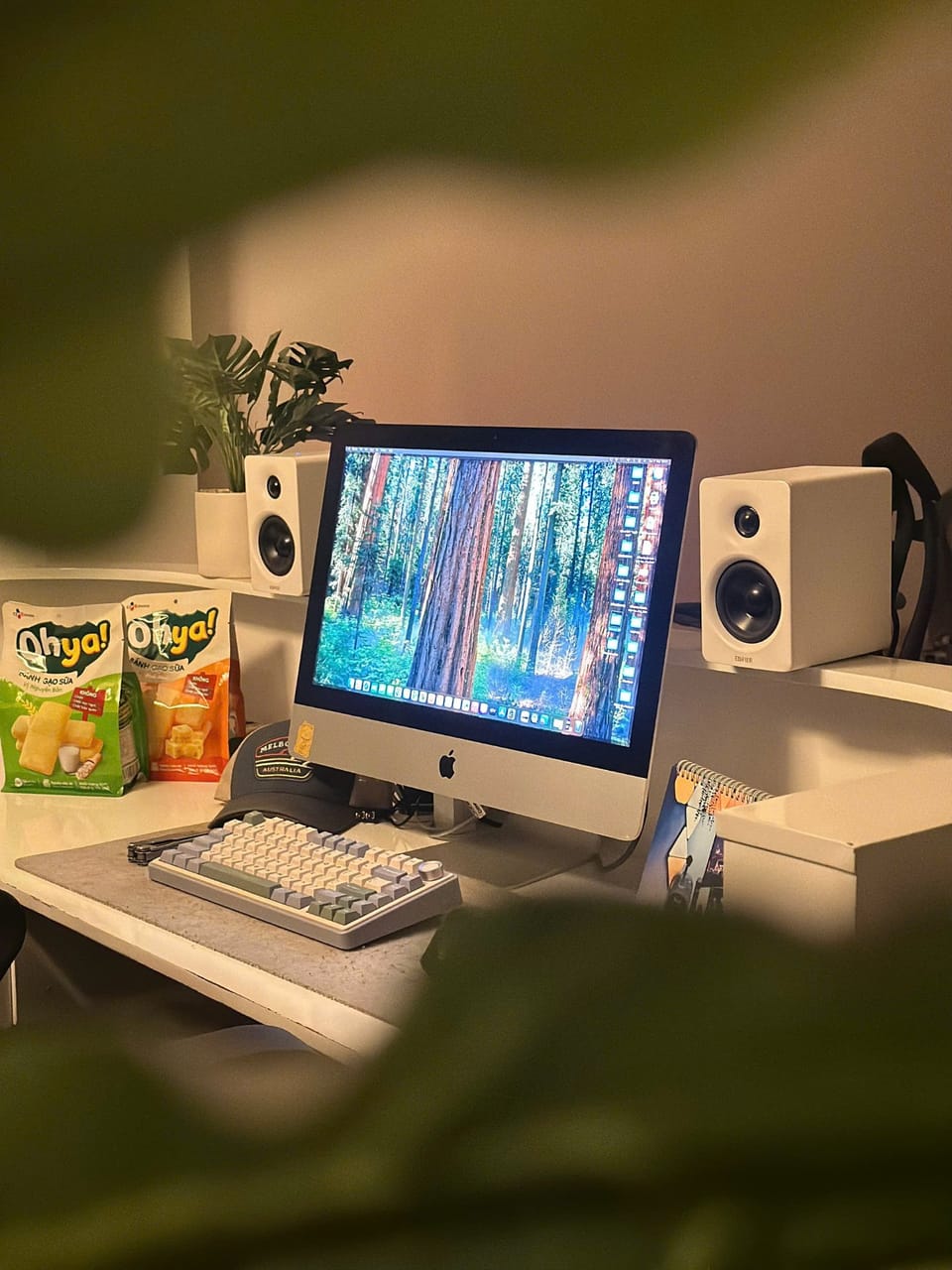 Góc máy của TheManson: Góc làm việc tông trắng với iMac, bàn phím cơ phối màu, cặp loa trắng, chậu cây xanh và vài gói bánh Ohya trên bàn.