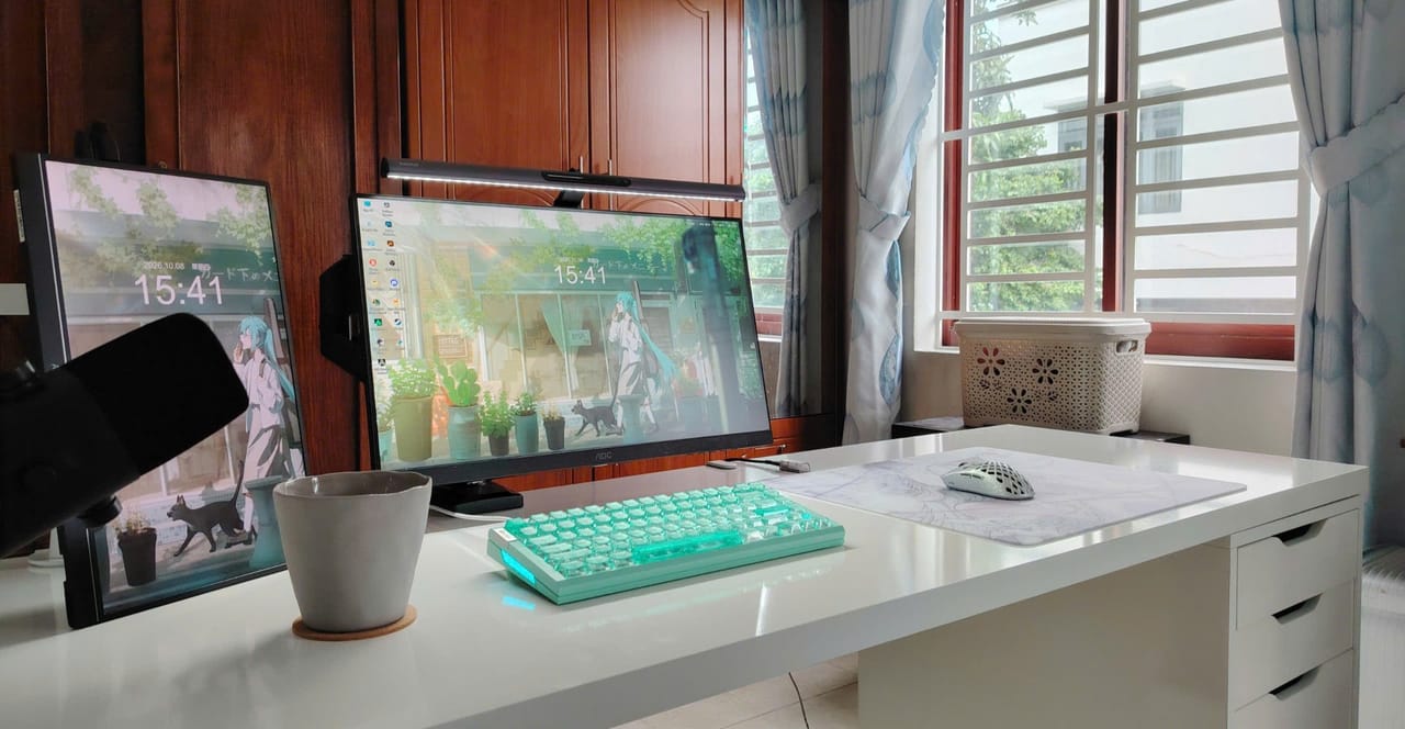 Góc máy của Zéphyr: Góc bàn trắng gọn gàng cạnh cửa sổ, có hai màn hình hình nền anime, bàn phím cơ xanh mint, chuột trắng và đèn treo màn hình.