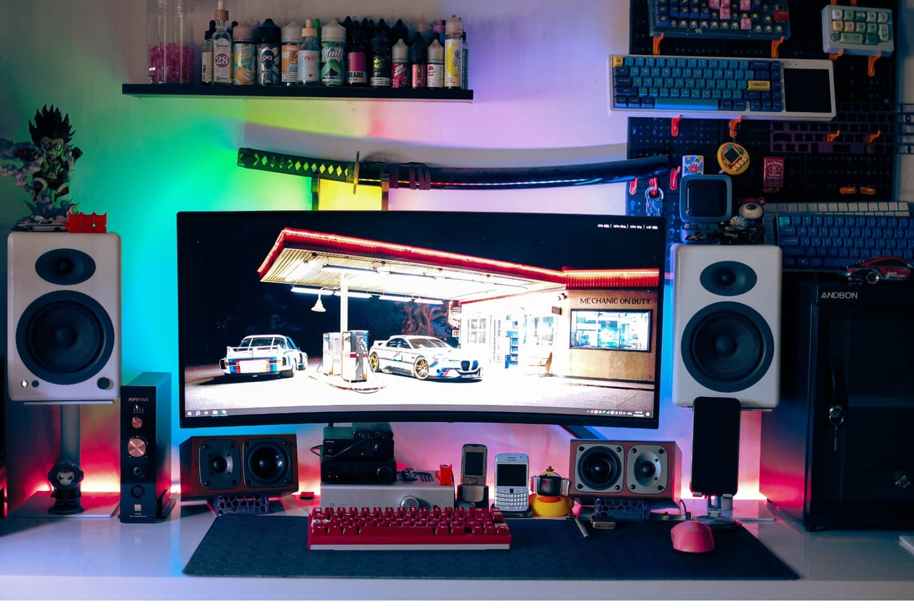Setup góc máy của nhocdkj: Góc máy với màn hình cong ultrawide, loa trắng hai bên, bàn phím đỏ, tường đèn RGB nhiều màu và kệ trưng bày bàn phím cùng mô hình.