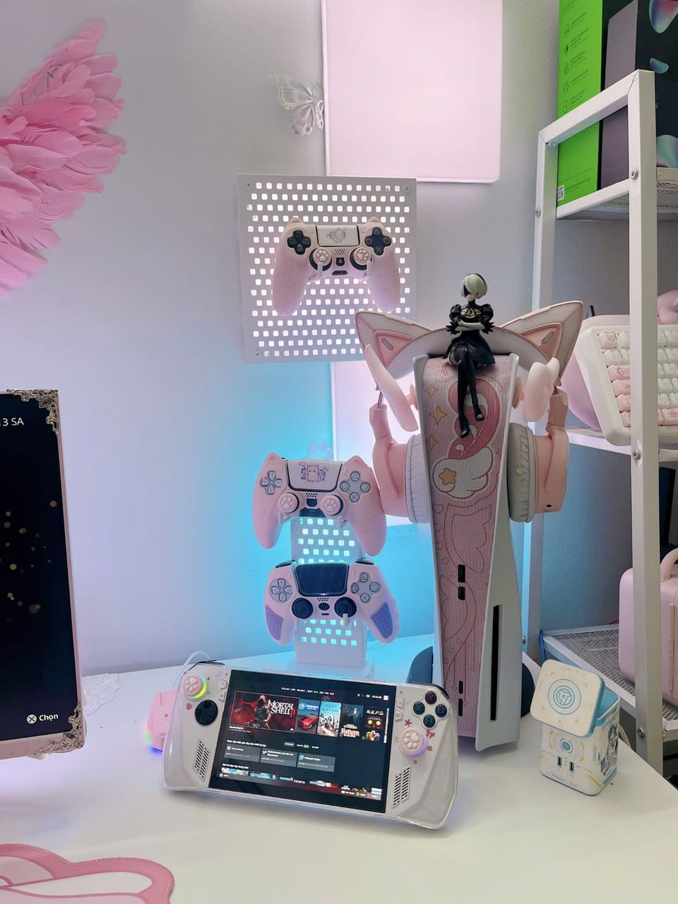 Góc máy của Dánh Gaming: Góc console tông hồng pastel với PS5 dán skin, tay cầm treo tường, tai nghe tai mèo, figure anime và máy chơi game cầm tay.