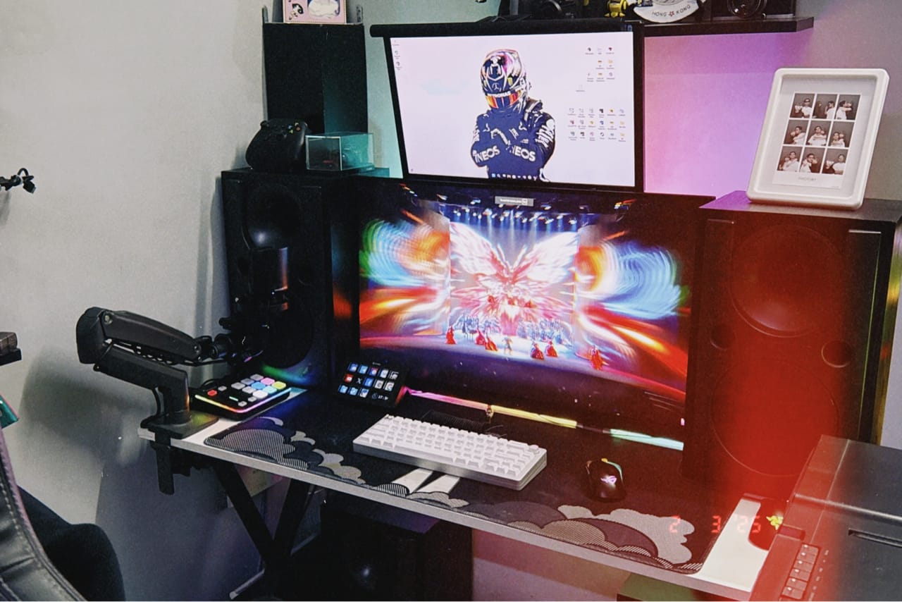 Góc máy của Khang Nguyen: Góc setup streamer với hai màn hình, loa lớn hai bên, bàn phím trắng, đèn RGB và khung ảnh photobooth trang trí.
