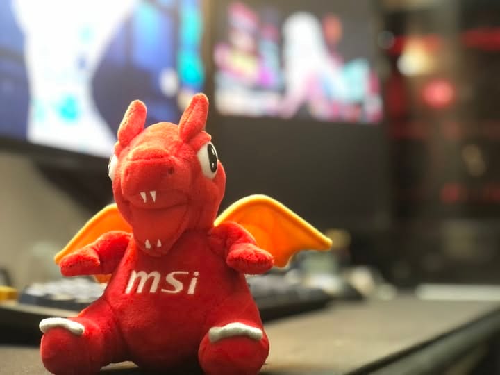 Góc máy của 0racle: Chú rồng nhồi bông đỏ logo MSI đặt trên bàn, phía sau là hai màn hình và bàn phím bị mờ.