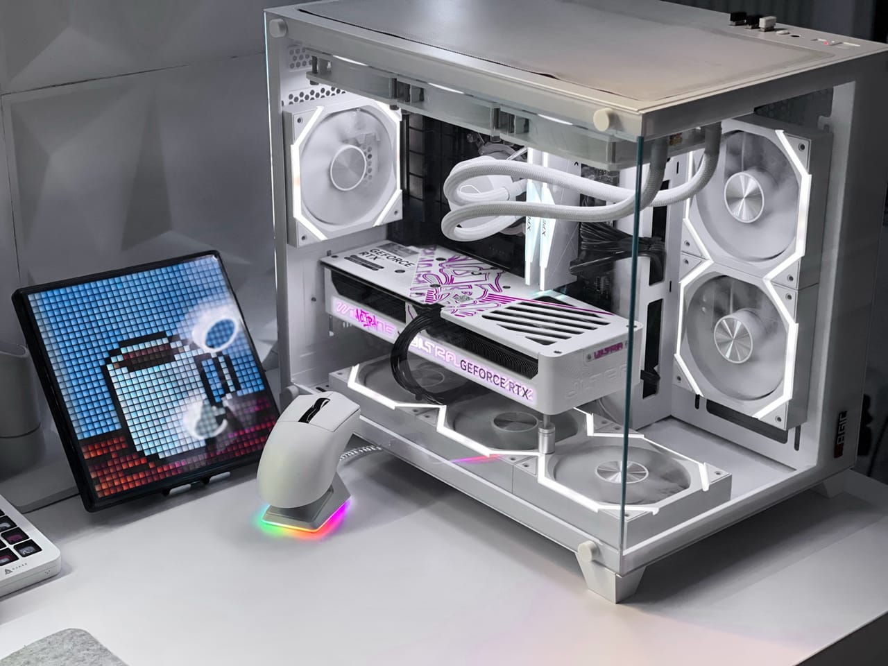 Góc máy của KhoaSetup: Case PC kính trắng với quạt viền LED trắng, card RTX trắng, chuột đế sạc RGB và màn hình pixel art trên bàn trắng.