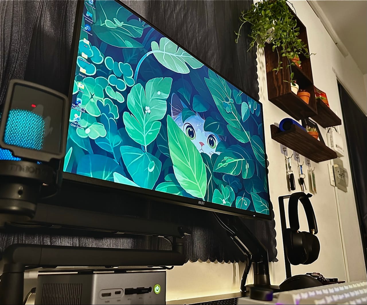 Góc máy của Võ Minh Sang: Góc máy tông đen với màn hình MSI hình nền lá xanh và chú mèo, mini PC, micro đèn xanh, tai nghe và kệ gỗ treo cây.