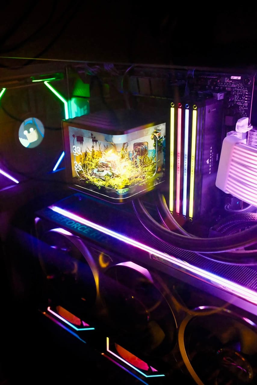 Góc máy của Tùng Dơi: Bên trong case PC với card đồ họa GeForce RTX, RAM Corsair Dominator sáng RGB và màn hình mini hiển thị tiểu cảnh cây xanh, ánh đèn neon tím vàng.