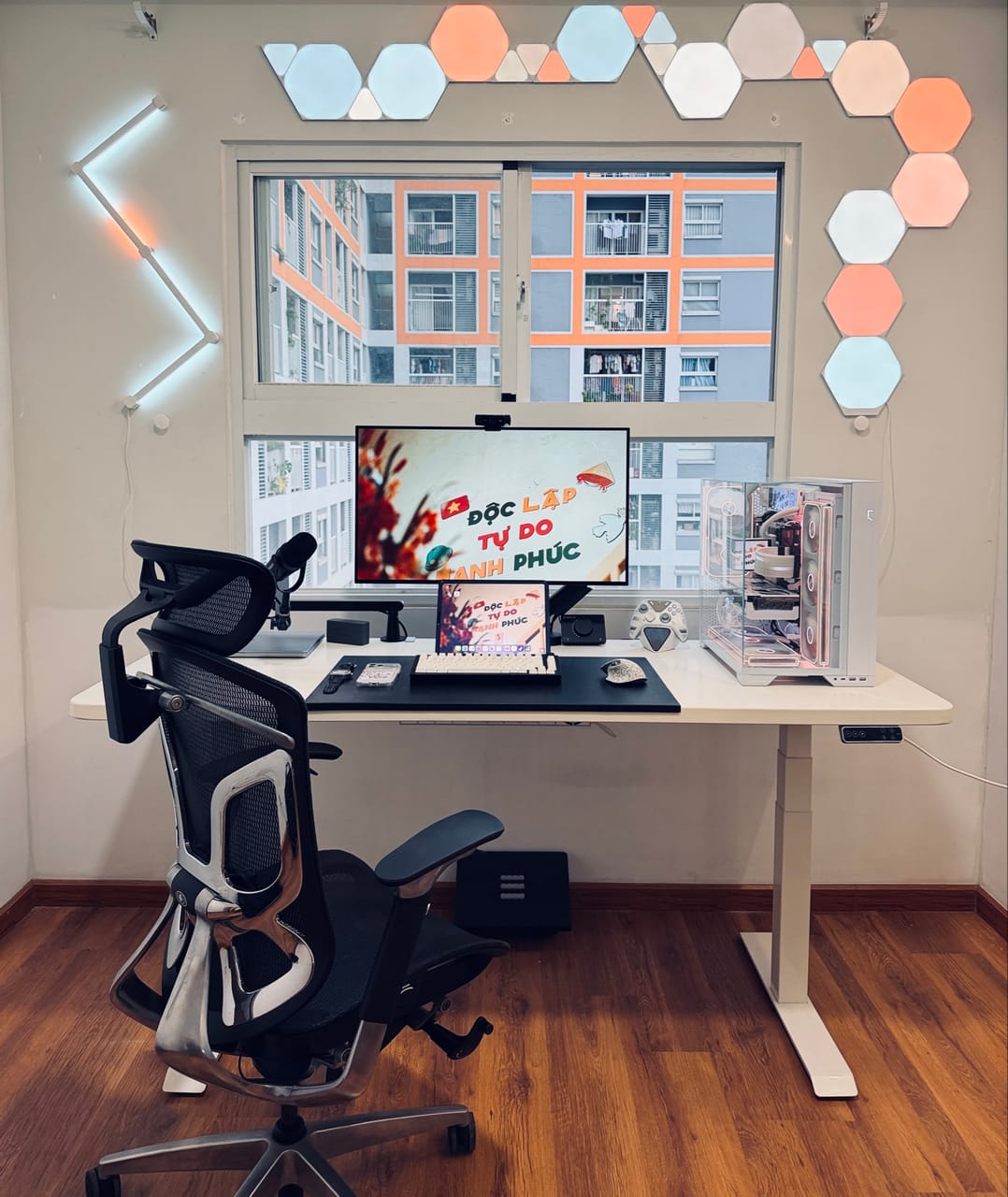 Góc máy của A giáo: Góc máy tông trắng với bàn nâng hạ, case PC kính trong suốt, màn hình, bàn phím cơ, ghế công thái học và đèn ốp tường hình lục giác nhiều màu.