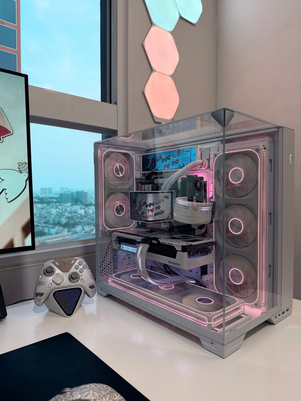 Góc máy của A giáo: Dàn PC vỏ kính trắng với đèn LED hồng pastel, card Radeon, tay cầm bạc và đèn lục giác trang trí cạnh cửa sổ.