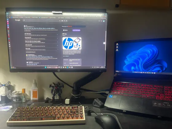 Góc máy của Kwan: Góc máy tông đen với màn hình HP trên arm, laptop Acer Nitro 5 phím đỏ, bàn phím cơ keycap cam và mô hình Gundam.