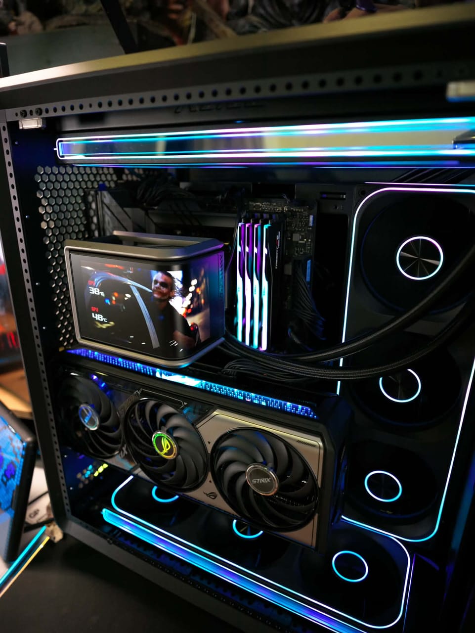 Góc máy của Tùng Dơi: Bên trong case PC kính với đèn LED RGB xanh tím, card đồ họa ROG Strix, RAM RGB, tản nhiệt nước và màn hình phụ nhỏ.