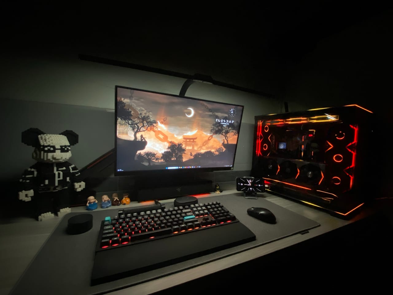 Góc máy của penguin: Góc máy tối với case PC viền LED cam đỏ, bàn phím đèn nền, màn hình hình nền hoàng hôn và mô hình gấu xếp hình bên cạnh.