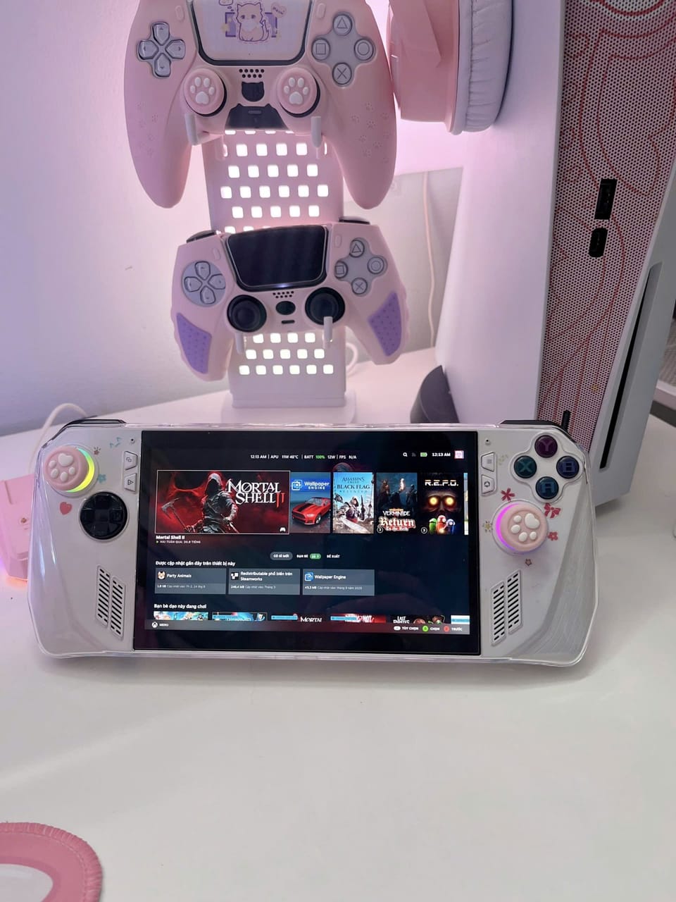 Góc máy của Dánh Gaming: Góc setup tông hồng pastel dễ thương với máy chơi game cầm tay, hai tay cầm gắn giá đỡ phát sáng, tai nghe và case trắng.