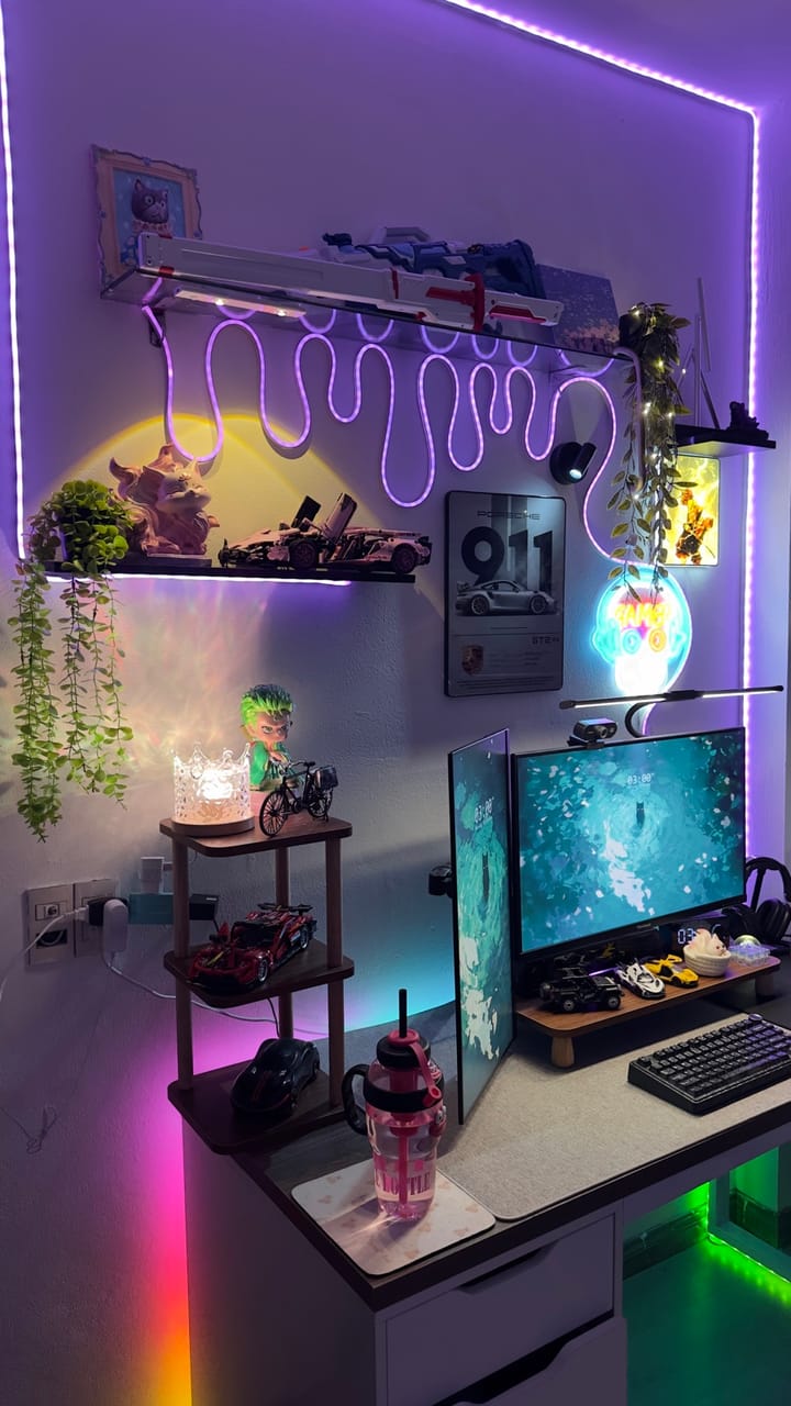 Góc máy của Kaii: Góc máy RGB tím rực rỡ với hai màn hình, đèn neon uốn lượn, mô hình siêu xe, figure anime và cây xanh trang trí.