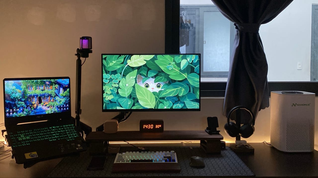 Góc máy của Võ Minh Sang: Góc máy tông đen và gỗ với màn hình hình nền lá xanh, laptop ASUS phím RGB, bàn phím cơ, micro và máy lọc không khí bên cửa sổ.