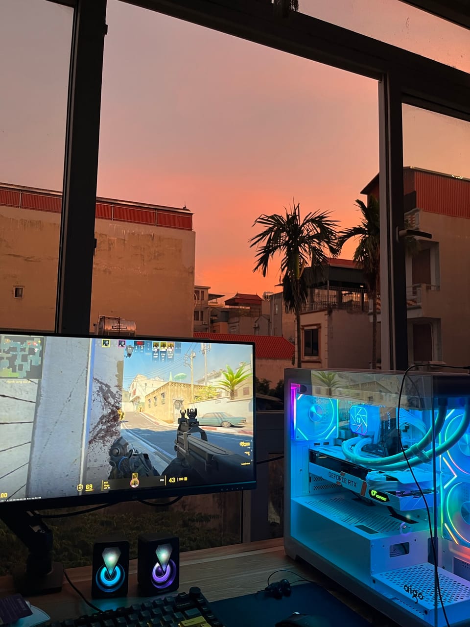 Góc máy của aug shadcn: Góc máy trắng RGB với case aigo tản nước, card RTX, chơi CS2 bên cửa sổ ngắm hoàng hôn rực cam.