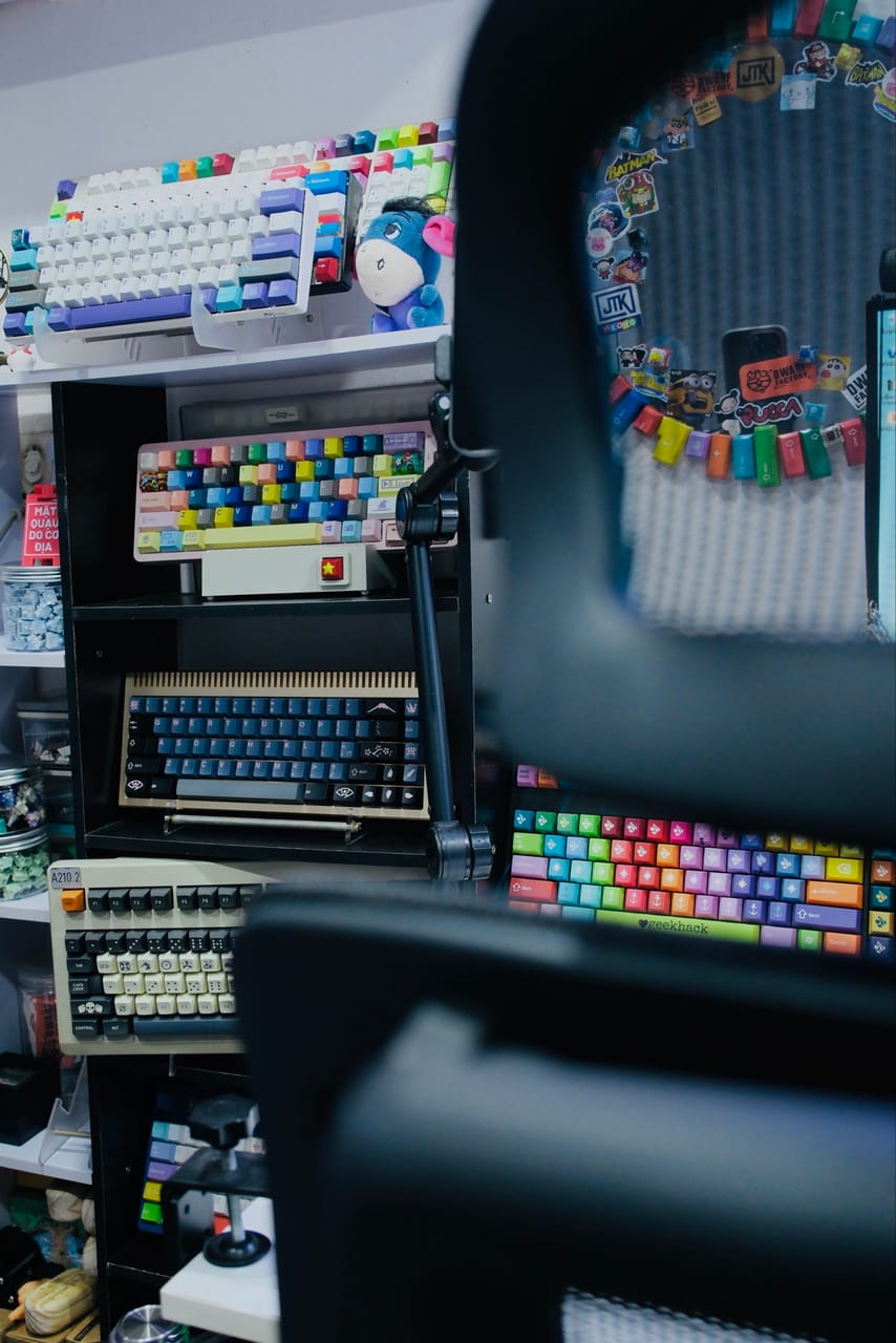 Góc máy của Thư Đoàn: Kệ trưng bày bộ sưu tập bàn phím cơ nhiều màu sắc, keycap rực rỡ, có arm màn hình và ghế nhìn ở phía trước.