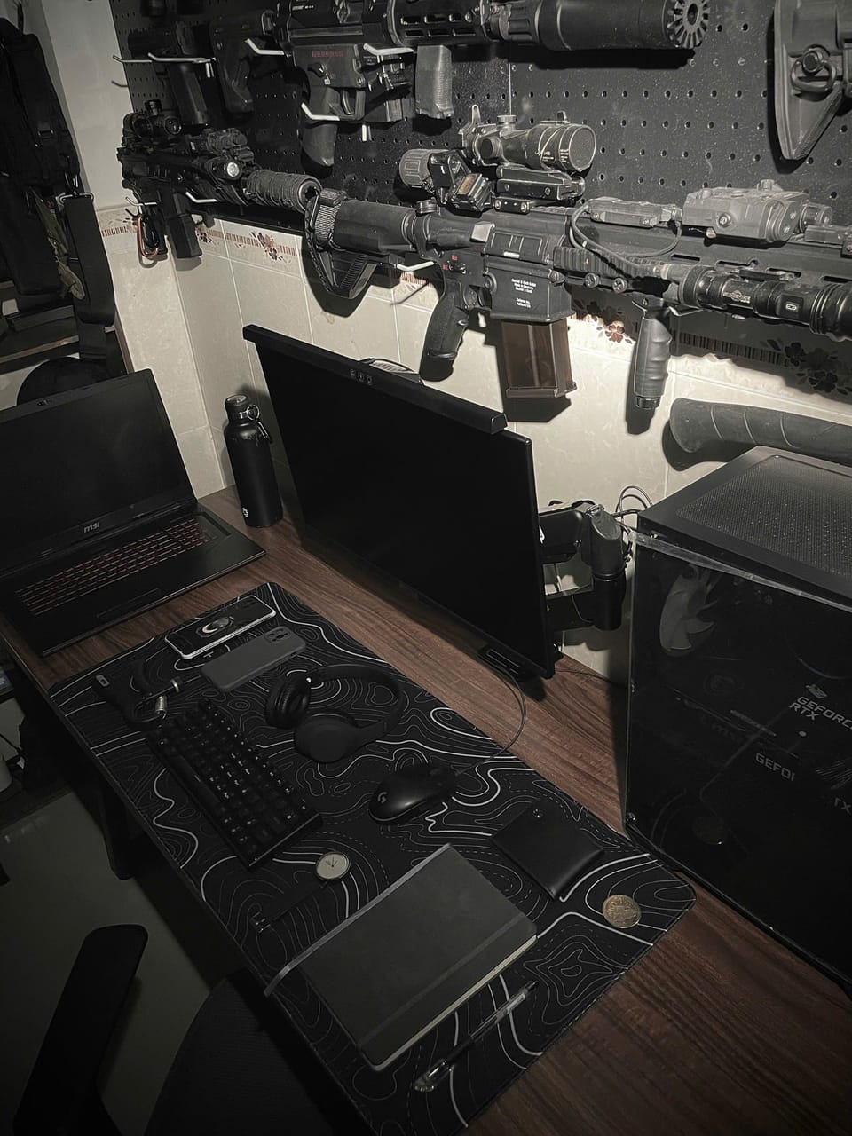 Góc máy của Harip: Góc máy tông đen tối giản với màn hình gắn arm, laptop MSI, case PC và lót chuột họa tiết địa hình trên bàn gỗ nâu.