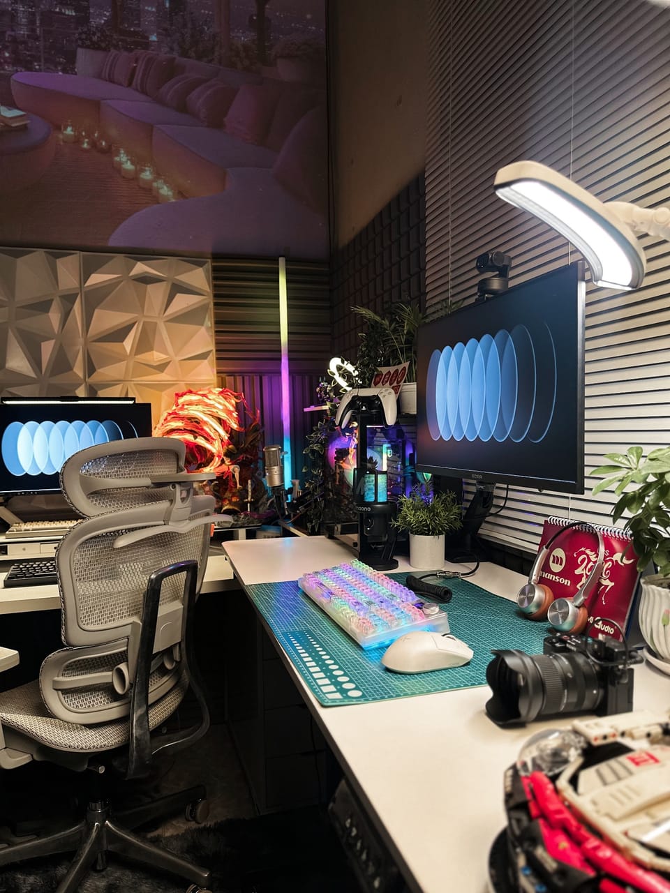 Góc máy của Drake nè: Góc setup đèn RGB nhiều màu với hai màn hình, bàn phím trong suốt, chuột trắng, mic, webcam, máy ảnh, cây xanh và ghế công thái học.