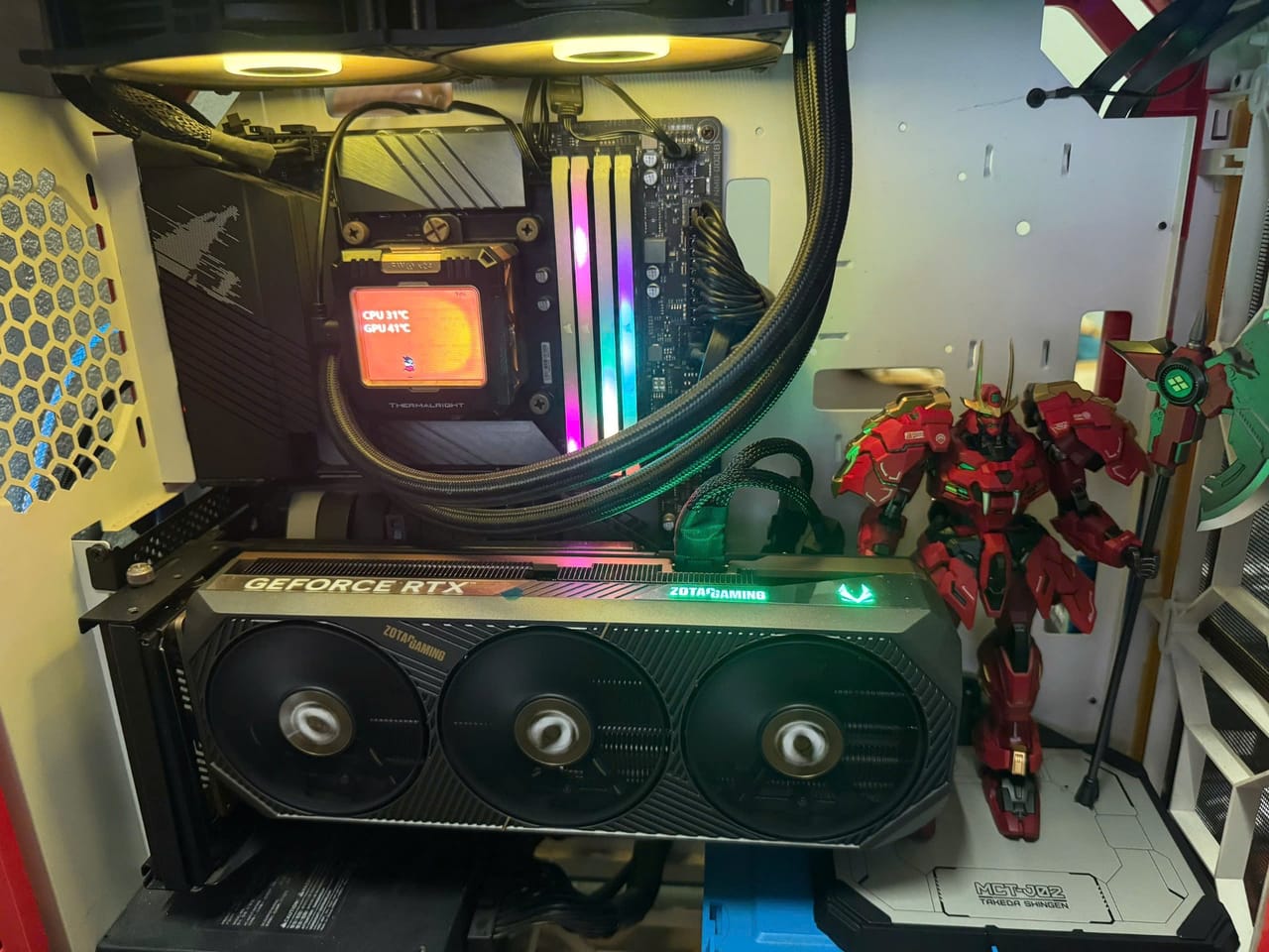 Góc máy của Shoa6: Bên trong case trắng đen có card đồ họa Zotac RTX, tản nhiệt nước Thermalright, RAM RGB và mô hình robot đỏ đặt cạnh.