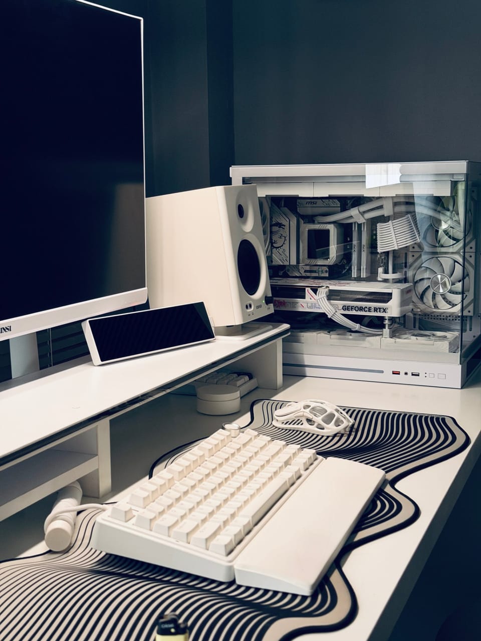 Góc máy của Chef Châu Tống: Góc setup tông trắng tối giản với PC kính trong suốt, bàn phím trắng, chuột lỗ tổ ong, loa và lót chuột họa tiết sóng đen trắng.