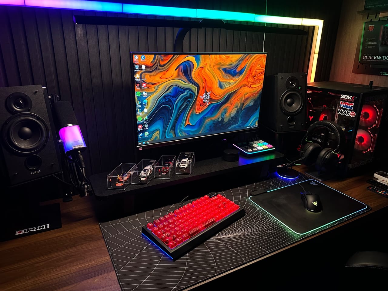 Setup góc máy của supra27: Góc setup tối với đèn RGB rực rỡ, bàn phím keycap đỏ trong suốt, màn hình hình nền Razer, loa Edifier và dàn mô hình xe.