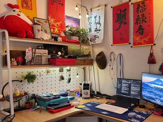 Góc máy của Long Bi: Góc làm việc bàn gỗ chữ L, laptop kèm màn hình rời, pegboard trắng, trang trí câu đối đỏ đậm chất Tết.