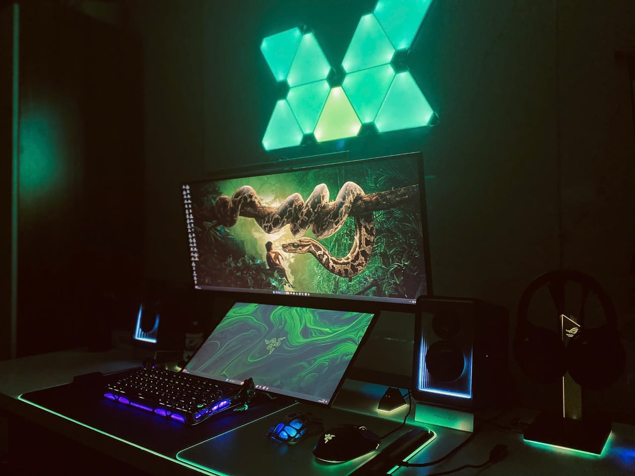 Góc máy của Tony Quân: Góc setup tối với đèn panel tam giác xanh lá trên tường, màn hình ultrawide, bàn phím, chuột Razer, loa và tai nghe ROG.