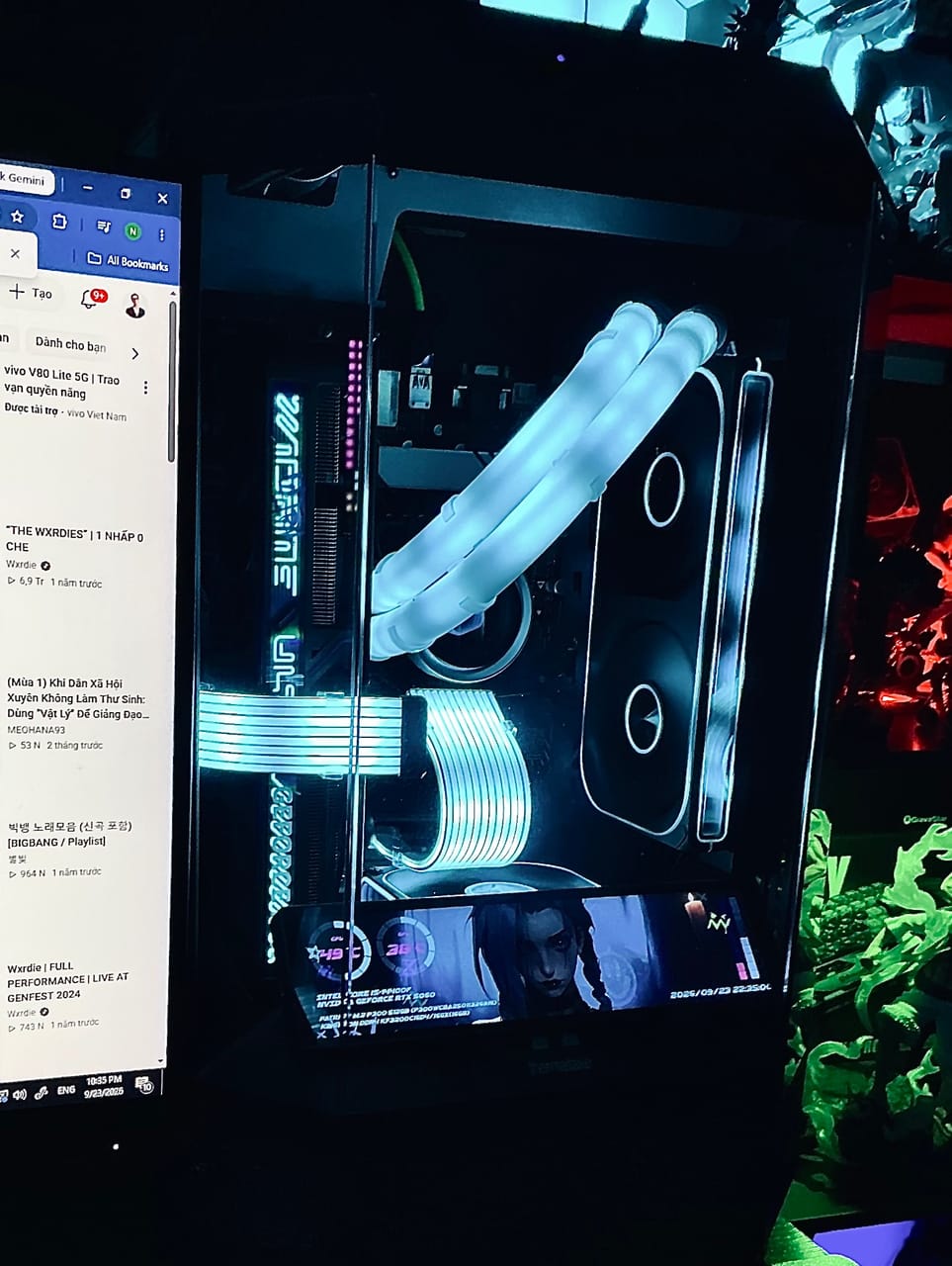 Góc máy của Rookie Bùi: Case PC kính trong suốt sáng đèn LED xanh trắng, có màn hình phụ hiển thị thông số, bên cạnh là mô hình figure xanh lá và đỏ.