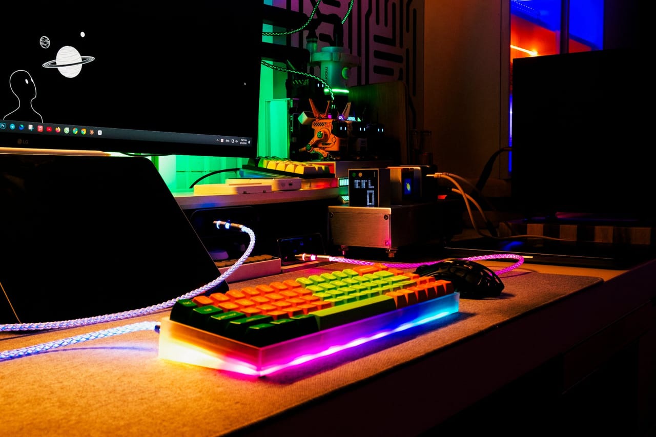 Góc máy của VGSer 9950: Góc máy tối với đèn neon xanh lá, tím hồng; bàn phím 60% keycap cam xanh viền acrylic RGB, chuột lỗ tổ ong và dây cáp phát sáng.