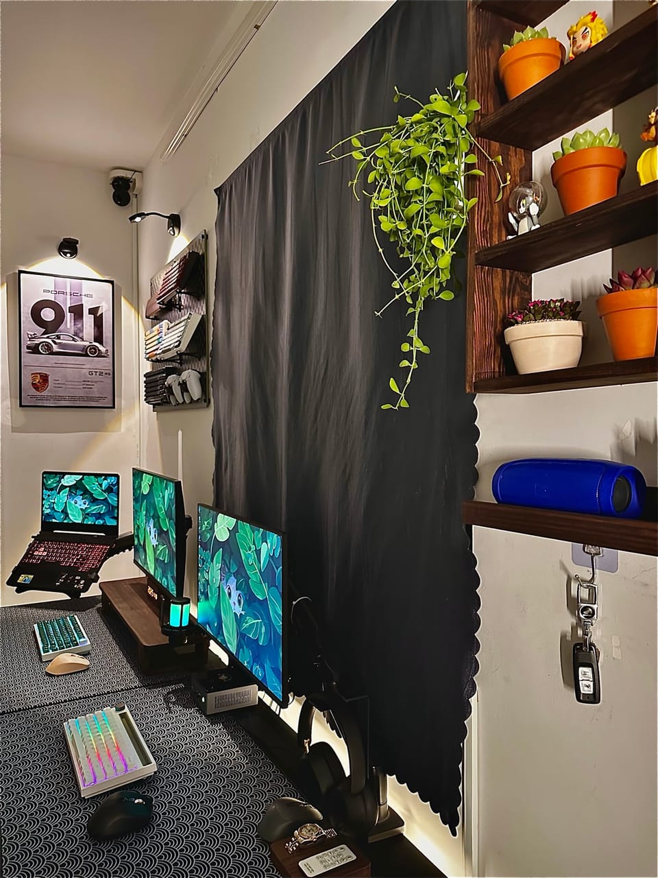 Góc máy của Võ Minh Sang: Góc setup nhỏ gọn với hai màn hình, laptop trên giá đỡ, kệ gỗ đặt sen đá, rèm đen và tranh Porsche 911 trên tường.