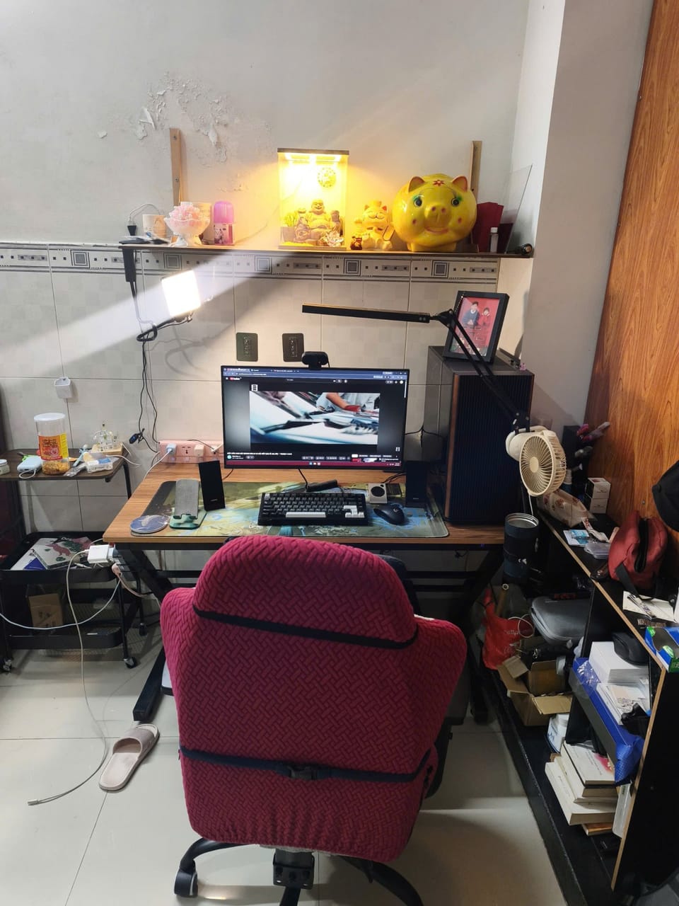 Góc máy của QuangPhu: Góc làm việc nhỏ với bàn gỗ, một màn hình, bàn phím cơ, đèn livestream, kệ thờ tượng Phật và heo đất vàng phía trên.