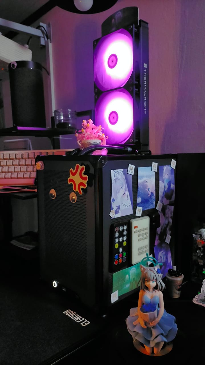 Góc máy của Tùng Lem: Case PC màu đen đặt tản nhiệt Thermalright phát sáng RGB tím, dán nhiều postcard anime, bên cạnh có figure nhân vật anime váy xanh.