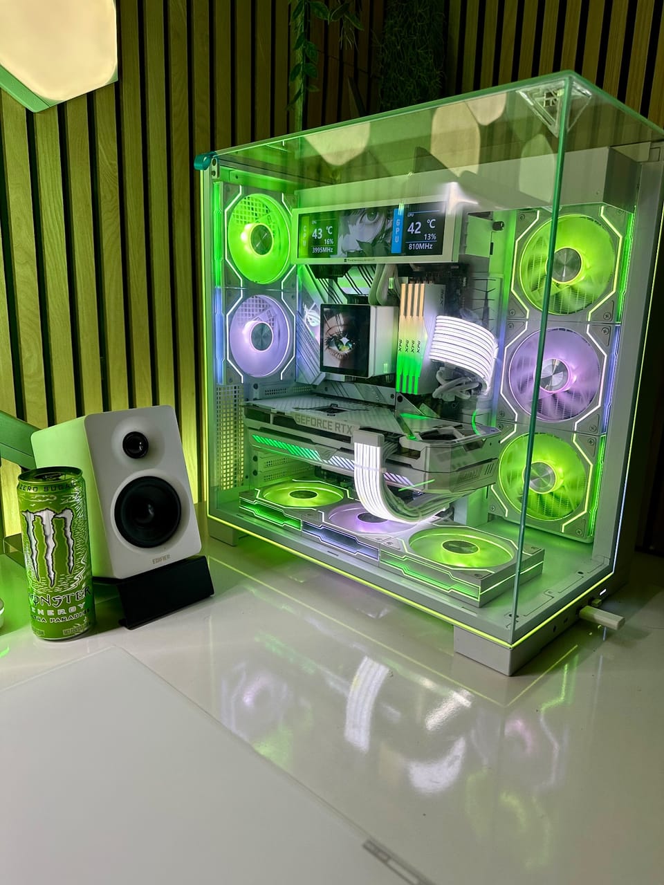 Góc máy của Quang Cozy: Case PC kính trắng với quạt RGB xanh lá và tím, cạnh loa Edifier trắng và lon Monster trên bàn, tường ốp gỗ nan.