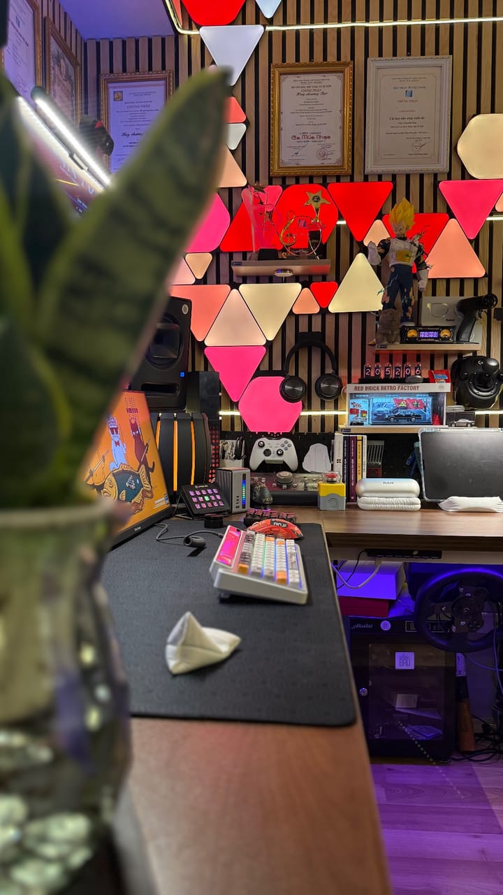Setup góc máy của Tùng Bầu: Góc setup gaming với đèn panel tam giác nhiều màu trên tường ốp gỗ, mô hình anime, bàn phím cơ, tay cầm và vô lăng chơi game.